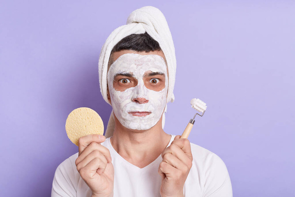 Image of shocked scared man with mask for skin, standing with towel on the head, holding sponge and massage roller for face, isolated over violet background. - Photo, Image