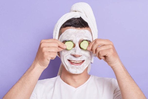 Indoor shot of smiling happy Caucasian man with mask for skin and covering eyes with cucumber slices standing wrapped in towel isolated over violet background. - Foto, afbeelding