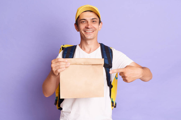 Horizontal shot of smiling satisfied delivery man holding pointing at paper packet with food isolated on violet background, looking smiling at camera. - Фото, зображення