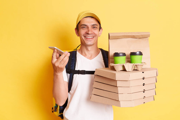 Portrait of happy delighted man in yellow cap and t-shirt, working as dealer courier, holding pizza and paper cups of coffee and mobile phone, posing isolated on yellow background. - Фото, зображення