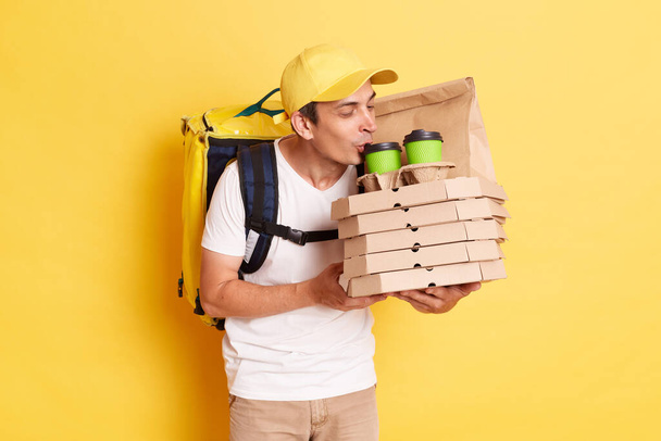 Portrait of joyful man in yellow cap and t-shirt, holding pizza and smelling coffee, likes his work, enjoying odor of oder, posing isolated on yellow background. - Foto, imagen
