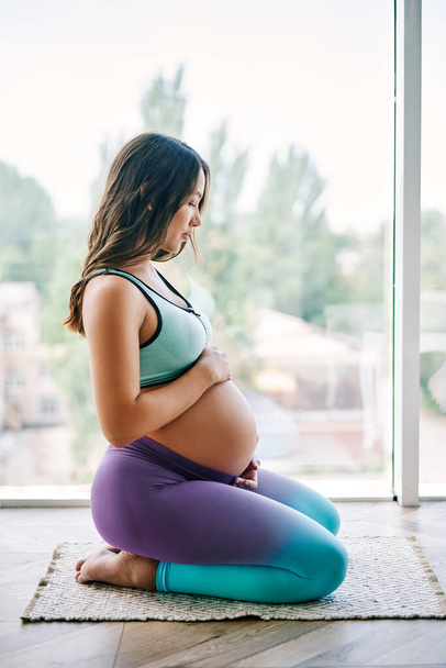 Portrait of young pregnant woman in sportswear sitting against big window holding hands on her belly. Pregnancy, expectation and tenderness concept. Conscious motherhood. - Fotó, kép