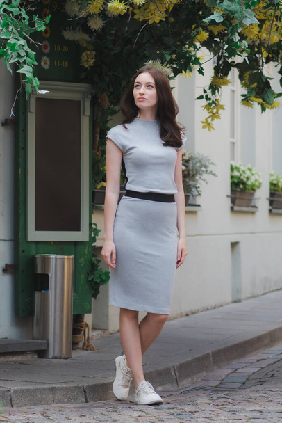 Piękna biała dziewczyna w krótkiej szarej sukience spaceruje ulicami starego miasta w pobliżu restauracji lub kawiarni z zieloną rośliną z kwiatami i tablicą na tle, przestrzeń do kopiowania, pionowe - Zdjęcie, obraz