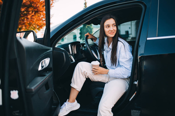 A happy attractive woman holding a cup of coffee and getting out of her modern car - Foto, immagini