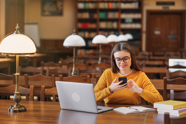 szczęśliwa studentka siedząca przy stole i czytająca SMS-y na swoim młodym telefonie. dziewczyna używa telefonu komórkowego w bibliotece koledżu - Zdjęcie, obraz