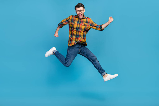 Full size photo of astonished delighted guy jumping raise fists triumph isolated on blue color background. - Foto, afbeelding