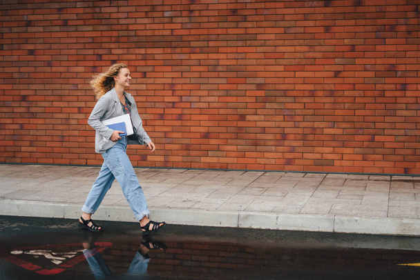Retrato de vista lateral de una estudiante que camina al aire libre contra el fondo de la pared de ladrillo, sosteniendo el portátil blanco mientras sonríe. Fondo urbano. Estilo de vida juvenil - Foto, imagen