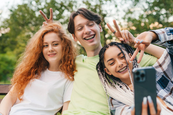 Edessä näkymä kolme ystävää istuu puinen penkki puistossa ottaen selfie käyttäen modernia laitetta. Selfie nuorten hymyilevien ihmisten hauskaa yhdessä - Valokuva, kuva