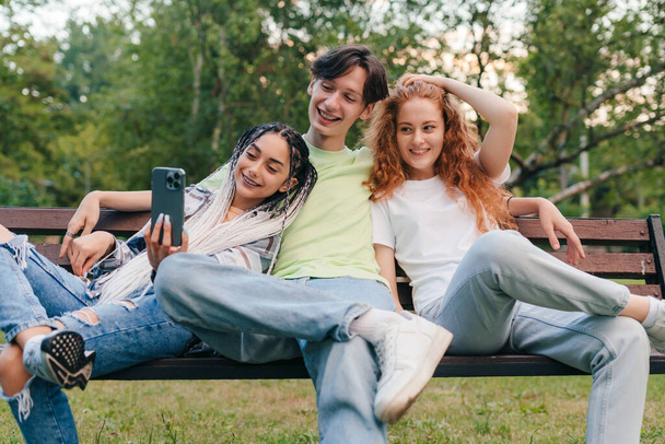 Τρεις έφηβοι blogger κάθονται σε ξύλινο πάγκο στο πάρκο το καλοκαίρι χρησιμοποιώντας ένα smartphone για την επικοινωνία βίντεο. Σύγχρονη επικοινωνία. Πράσινη πόλη. Απόσταση - Φωτογραφία, εικόνα