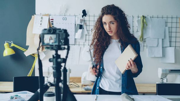 Young attractive seamstress fashion blogger is recording video with camera, talking and showing sketches and diferent tailoring items. Blogging for successful startup concept. - Fotoğraf, Görsel