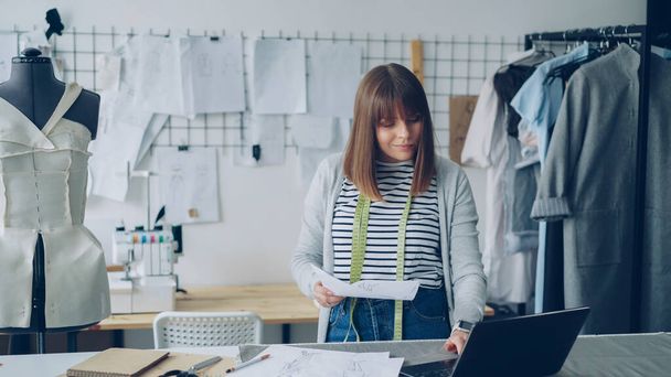 Young attractive woman is choosing sketches for new fashion show and using laptop in her modern tailors shop. Creative thinking in clothing design industry concept. - Photo, image