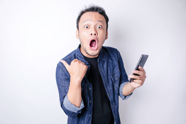 Surprised Asian man wearing navy blue shirt pointing at his smartphone, isolated by white background - Fotografie, Obrázek