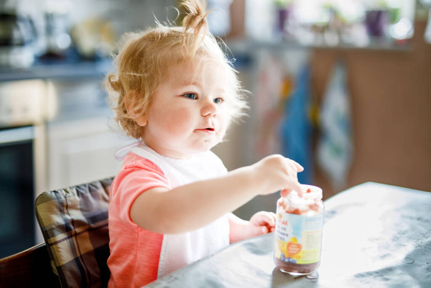 Urocza dziewczynka jedzenie z łyżką warzyw lub owoców konserwy, dziecko, karmienie i rozwój koncepcji. Słodkie małe dziecko, Córka z łyżką siedzi w krzesełko do karmienia i ucząc się jeść samodzielnie - Zdjęcie, obraz