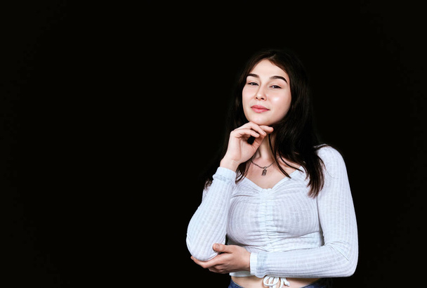 Beautiful young woman in white blouse resting her chin on her hand while looking at camera with satisfied emotion on her face on black background. Banner with copy space. Natural beauty, hair care, clear skin. - Photo, Image