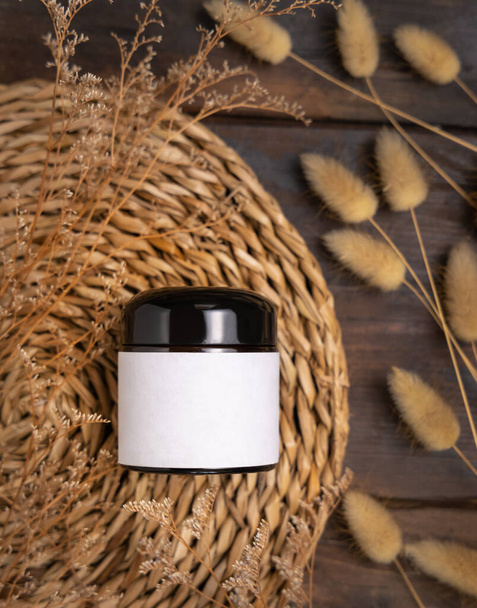 Cream jar on wattled boho placemat on wood near Hare's Tail grass top view, packaging label mockup. Natural skincare product for everyday beauty routine. Bohemian eco friendly cosmetics flat la - Фото, зображення