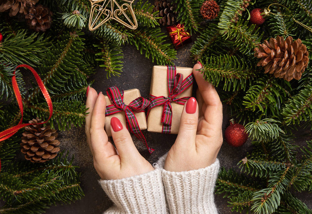 Female hands holding gift boxes near Christmas decorations and fir tree branches top view on a dark table. Christmas holiday atmosphere flat lay. Wrapping presents for loved ones during winter holidays - Photo, image