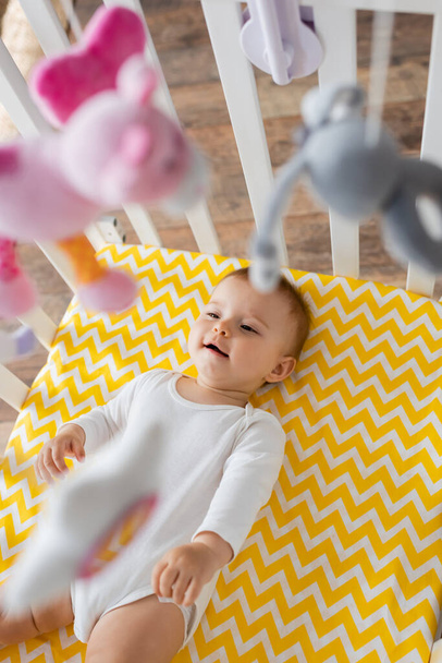 top view of happy infant girl in baby romper lying in crib and looking at hanging mobile with soft toys - Foto, Bild