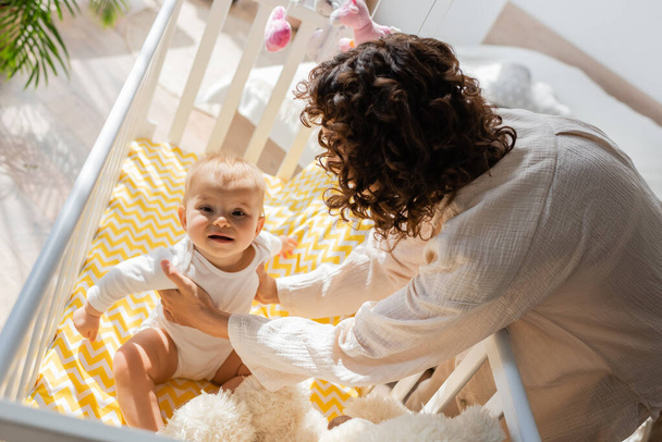 high angle view of curly mother in loungewear taking baby daughter in romper from crib  - Zdjęcie, obraz