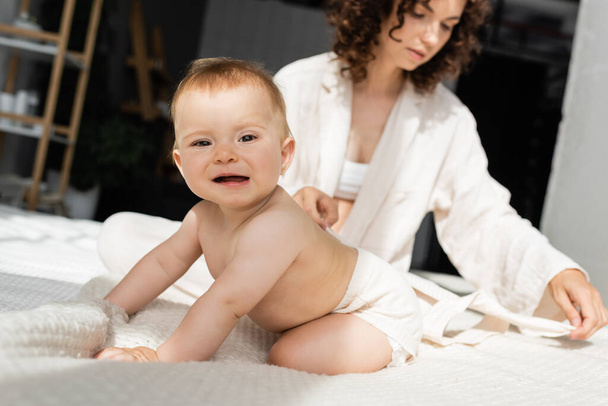 csecsemő baba romper ül az ágyon közelében elmosódott és göndör anya  - Fotó, kép
