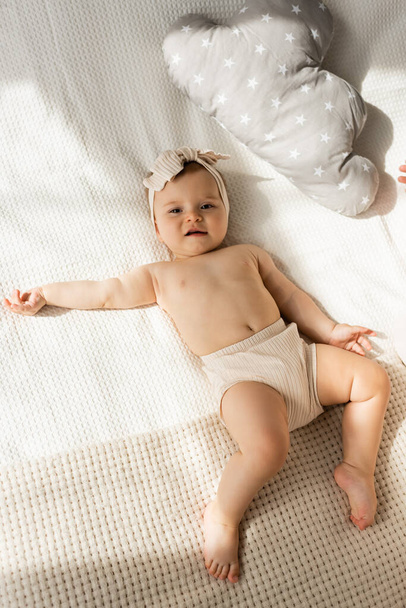 top view of barefoot infant girl in headband lying on bed near pillow in cloud shape  - Foto, imagen