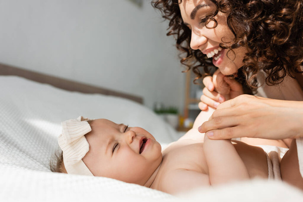 fröhliche Mutter mit lockigem Haar, die Hände eines kleinen Mädchens im Stirnband haltend  - Foto, Bild