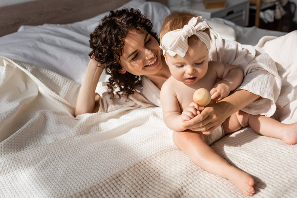 положительная женщина с кудрявыми волосами лежит на кровати с младенческой дочерью в головном уборе держа деревянную игрушку - Фото, изображение
