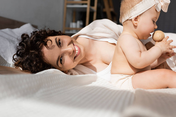mère heureuse couchée sur le lit et regardant la caméra près de bébé fille avec hochet en bois  - Photo, image