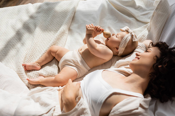 vue du dessus de la mère heureuse allongée sur le lit et regardant bébé fille dans le bandeau avec arc tenant hochet en bois  - Photo, image