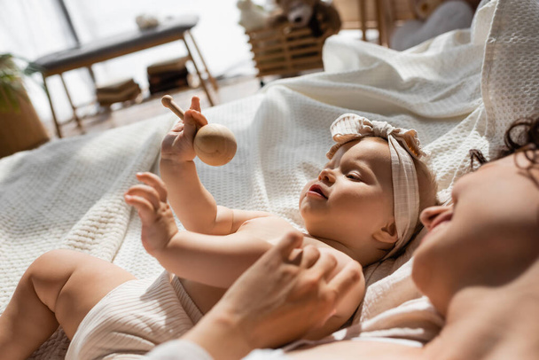 fröhliche Mutter liegt im Bett neben Baby-Mädchen mit Stirnband und hölzerner Rassel - Foto, Bild