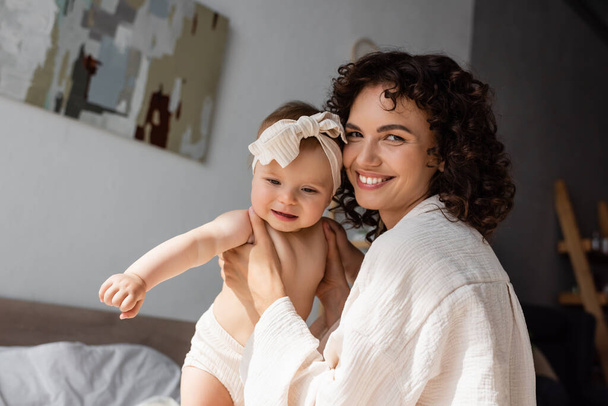 cheerful and curly mother holding in arms baby girl in headband with bow  - Foto, immagini