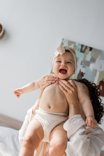 curly woman holding excited infant daughter in headband with bow and underwear  - Foto, Imagem