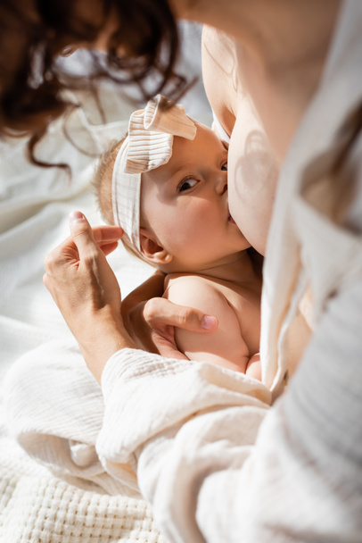 высокий угол зрения кудрявой женщины, кормящей грудью младенца, в головном уборе с бантом  - Фото, изображение