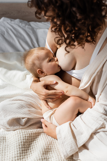 high angle view of curly mother in loungewear breastfeeding infant daughter - Фото, зображення