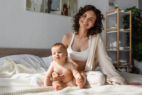cheerful mother in loungewear with crop top sitting near infant daughter in bedroom  - Fotó, kép