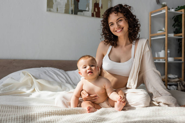 madre positiva en ropa de salón con la parte superior de la cosecha sentado cerca de la hija lactante en el dormitorio  - Foto, Imagen