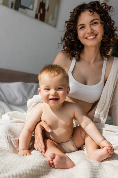joyful woman in loungewear with crop top sitting near infant daughter in bedroom  - Photo, Image