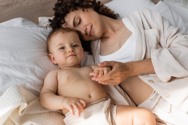 vue grand angle de la mère bouclée en vêtements de salon couché avec bébé fille sur le lit  - Photo, image