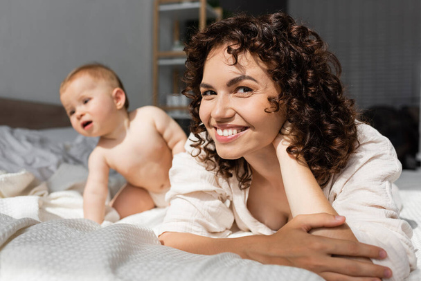 happy mother with curly hair looking at camera near blurred baby girl on bed - Фото, изображение