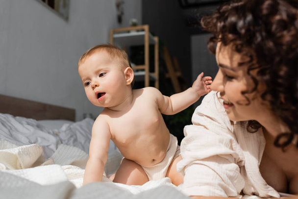 cute infant girl sitting on bed near blurred and curly mother  - Photo, Image