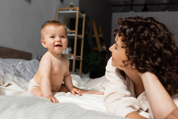 mother with curly hair looking at happy baby daughter sitting on bed - Foto, afbeelding