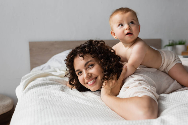 infant baby lying on back of happy mother with curly hair in bedroom  - Foto, immagini