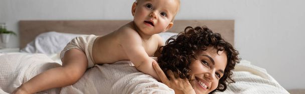 Säugling liegt auf dem Rücken einer glücklichen Mutter mit lockigem Haar im Schlafzimmer, Banner - Foto, Bild