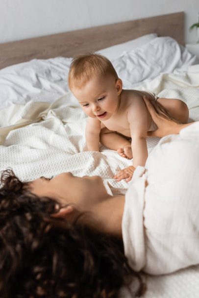 Säugling sitzt neben fröhlicher und lockiger Mutter auf dem Bett - Foto, Bild