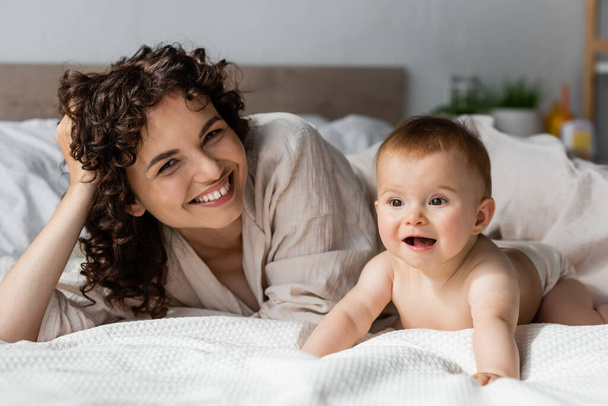 mujer feliz con el pelo rizado sonriendo mientras mira a la cámara cerca de bebé emocionado arrastrándose en la cama - Foto, imagen