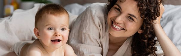 happy woman with curly hair smiling while looking at camera near excited infant baby, banner - Photo, Image