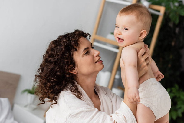 pleased woman with curly hair smiling while holding in arms excited baby daughter  - Photo, Image
