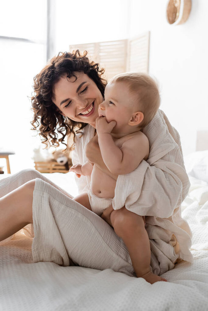 cheerful woman in loungewear holding in arms baby daughter and sitting on bed - Fotoğraf, Görsel