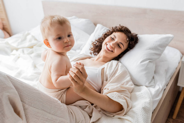 mujer feliz en ropa de salón acostada en la cama y cogida de la mano de la hija lactante  - Foto, imagen