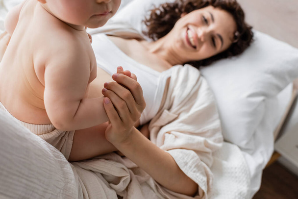 cheerful woman in loungewear lying on bed and holding hand of infant daughter  - Photo, Image