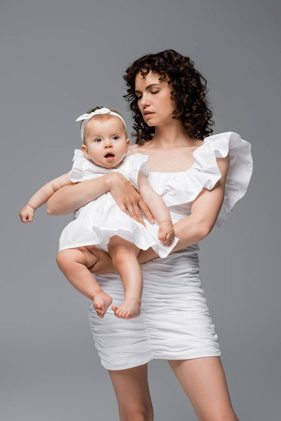 Stylish mother in white dress holding baby daughter isolated on grey  - Φωτογραφία, εικόνα